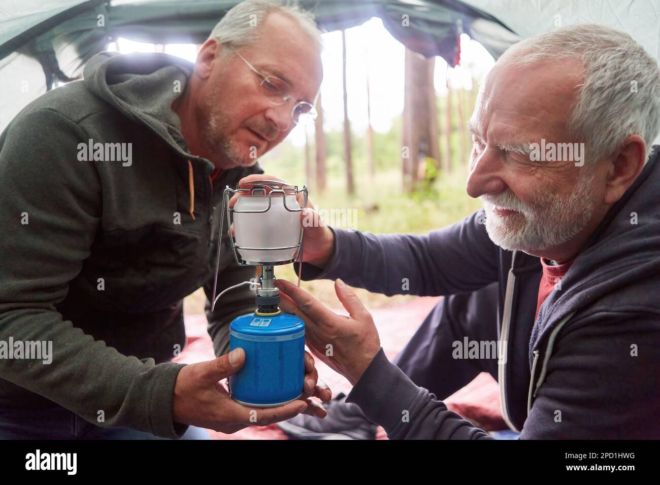 Elderly man fixing burner on stove with male friend while camping in tent during vacation Stock Photo