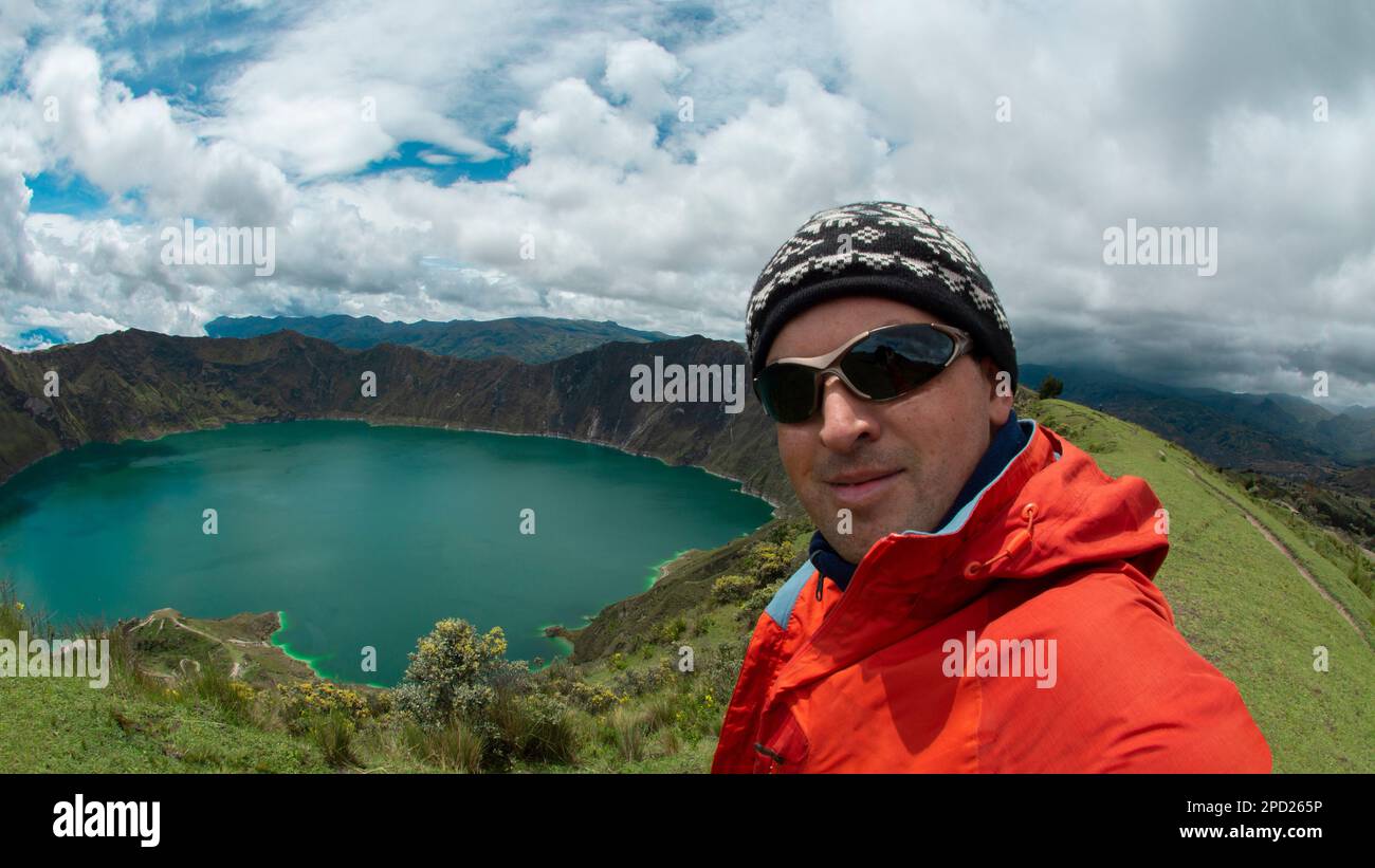 Young tourist man in red coat and glasses taking a selfie near the crater of Quilotoa volcano in Ecuador Stock Photo