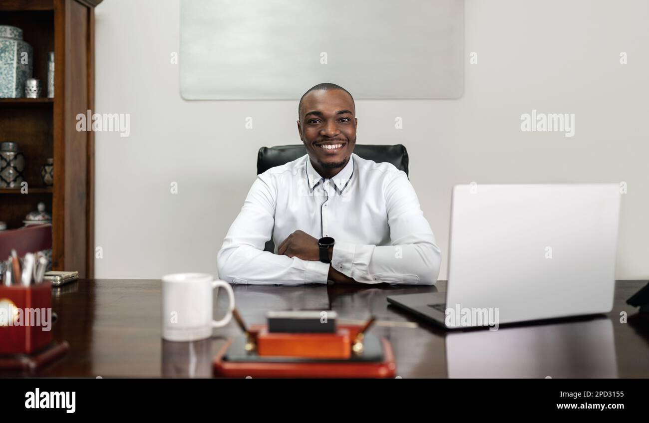 A mature African man sits at a table indoors, confidently crossing his arms as he uses his laptop for corporate business. Stock Photo
