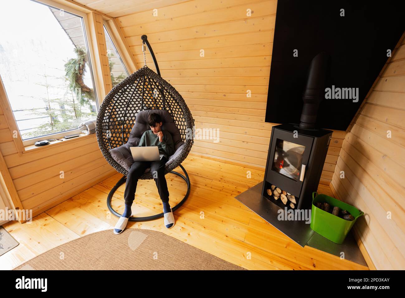 Teenager boy sit with laptop inside egg swing against fireplace at cozy indoors heating. Stock Photo