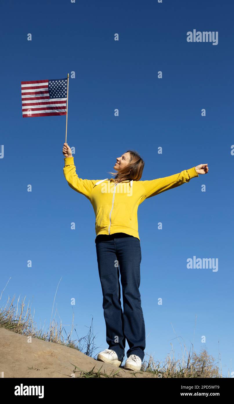 Patriotic happy woman joyfully raised up the American flag against a cloudless blue sky, 4th of July celebration. Independence Day of the United State Stock Photo
