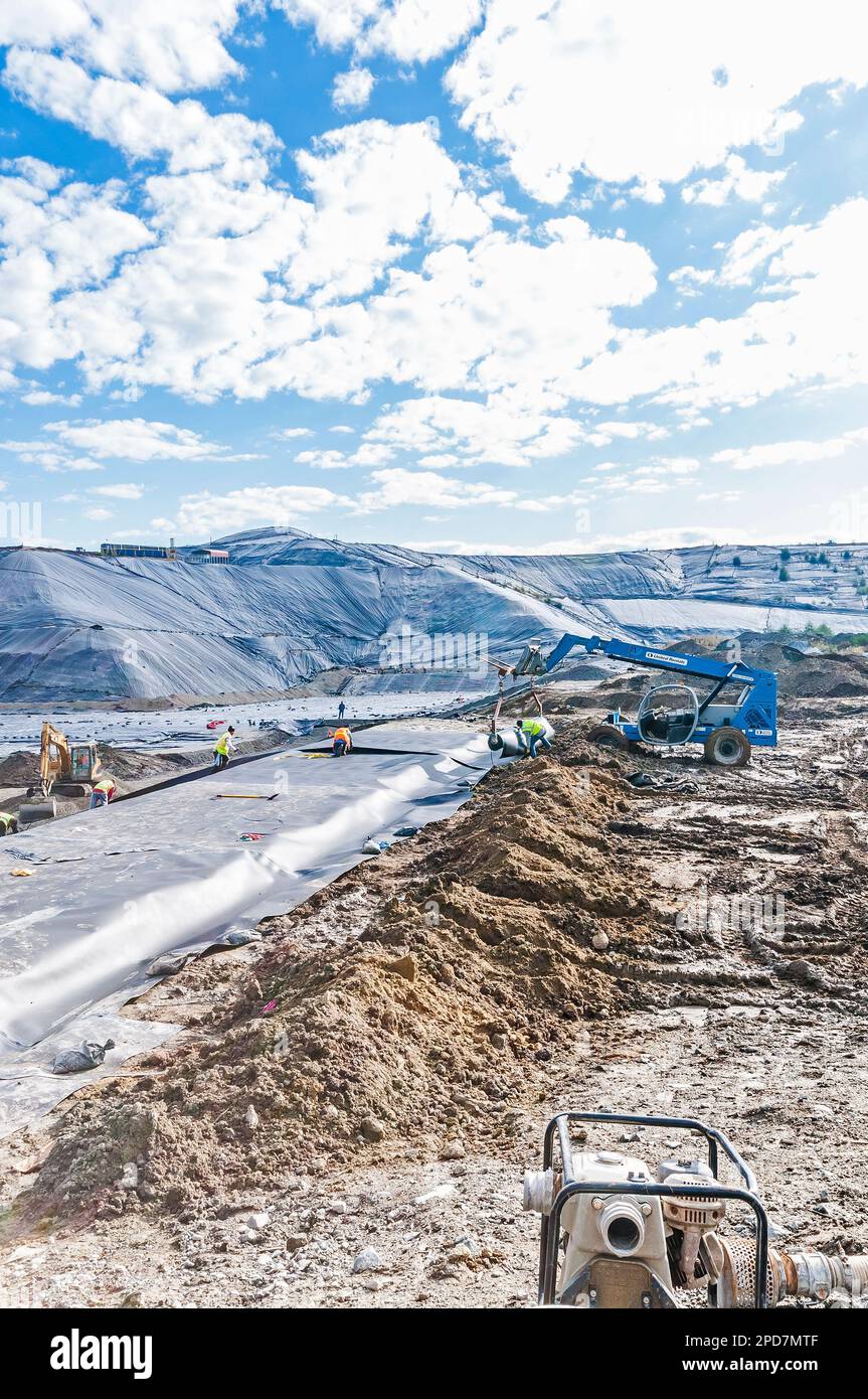 Workers with machinery are laying down plastic sheeting or geomembrane in an active landfill. Stock Photo