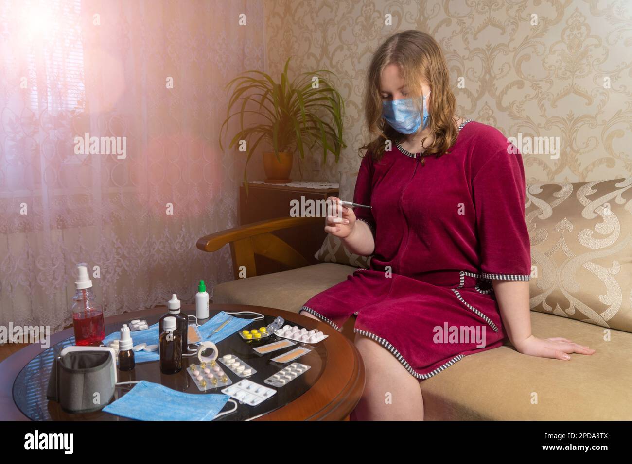 Girl in the room sits on the couch and checks her temperature on a thermometer. The concept of medicine, pharmacy, healthcare and treatment at home Stock Photo