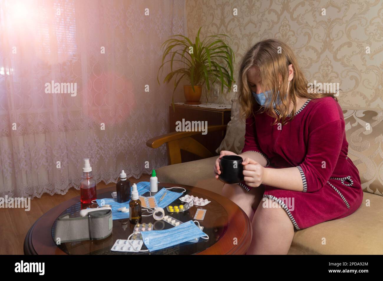 Girl with sits on a sofa in the room and holds a cup with a hot drink in her hands. Concept of medicine, pharmacy, healthcare and home treatment Stock Photo