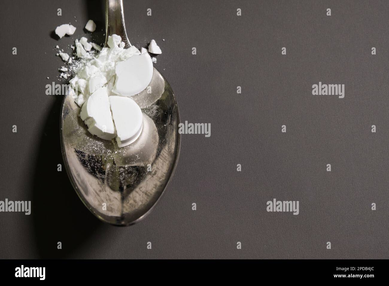 Pharmaceutical medicine drugs powder on a silver metal spoon. Smashed remedy, tablets, on top of a grey table in the background. Stock Photo