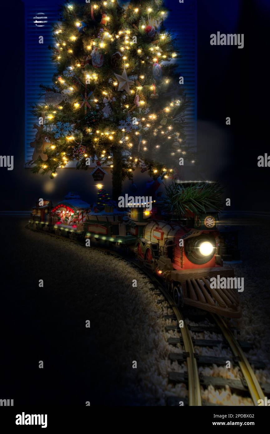 A toy model Christmas train on the floor below a decorated fake Christmas tree photoshopped to look like it’s at night Stock Photo