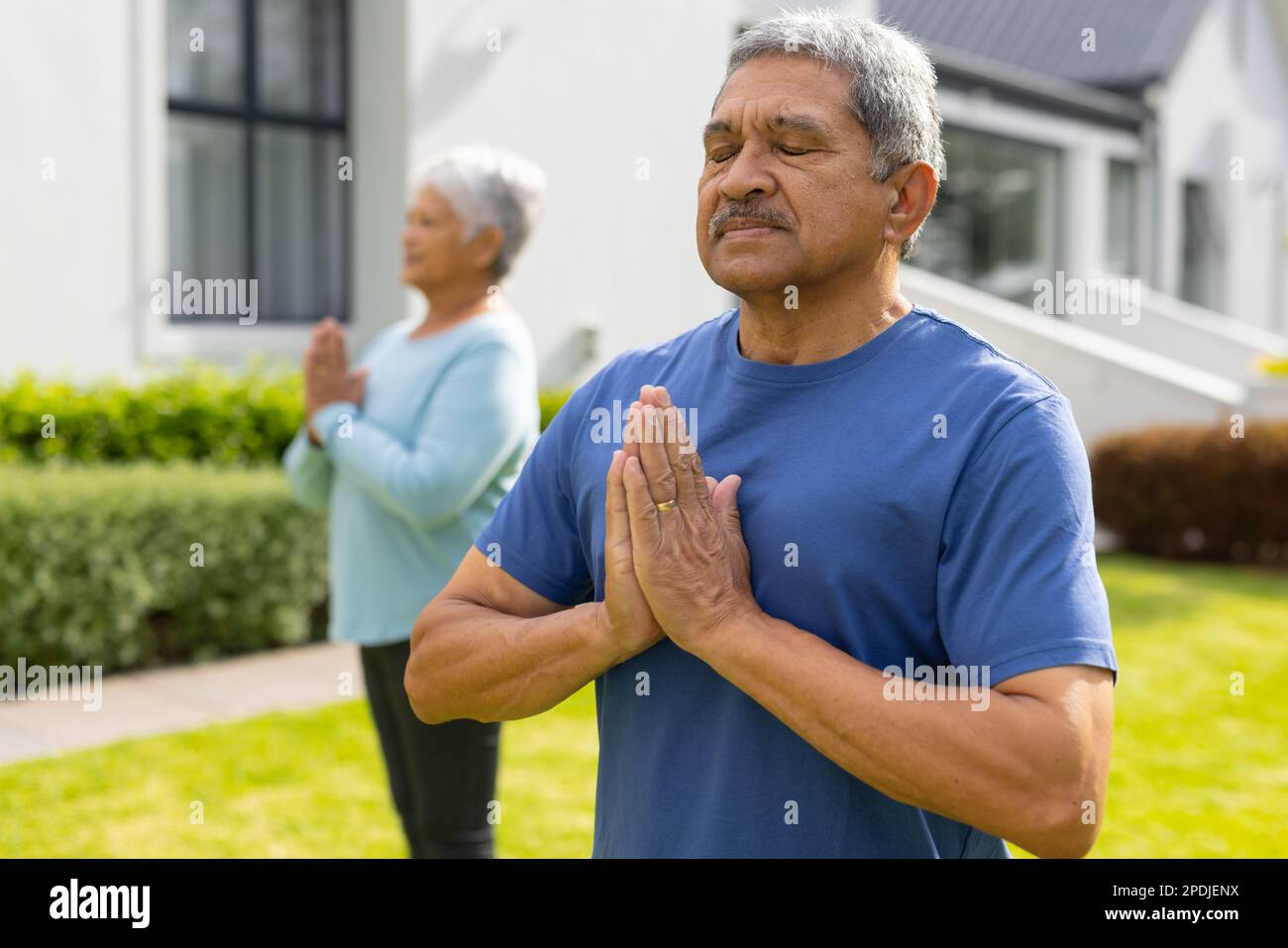 Biracial senior man and woman meditating in prayer pose against house at yard Stock Photo