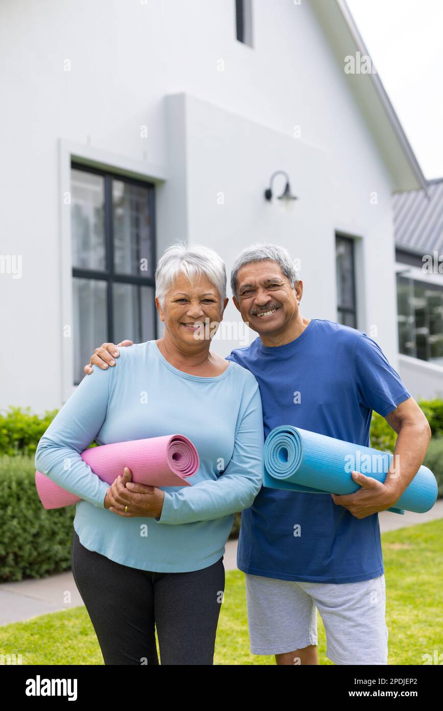 Portrait of smiling biracial senior couple with yoga mats standing against house in yard Stock Photo