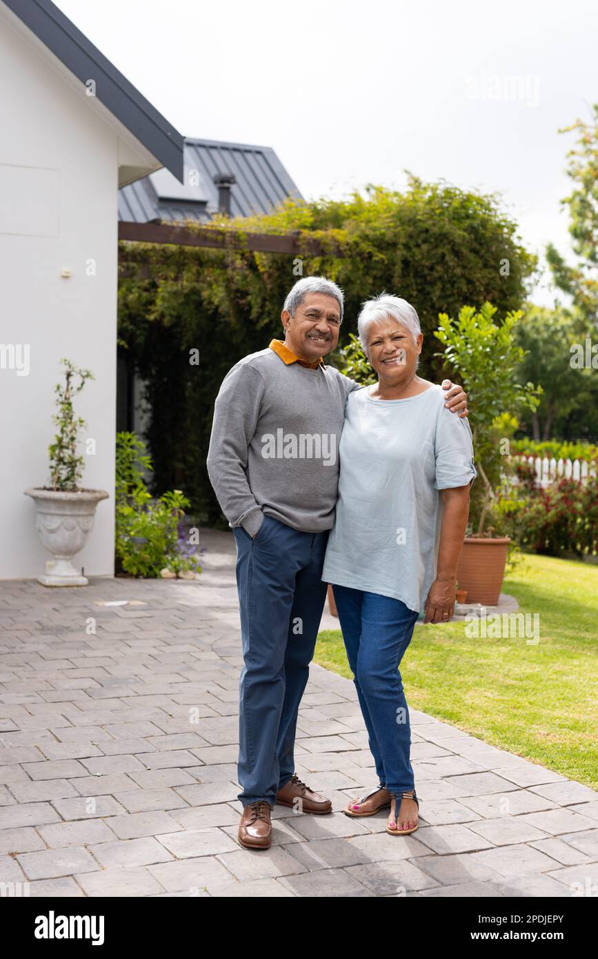 Portrait of smiling biracial senior man with hand on wife's shoulder standing on footpath in yard Stock Photo