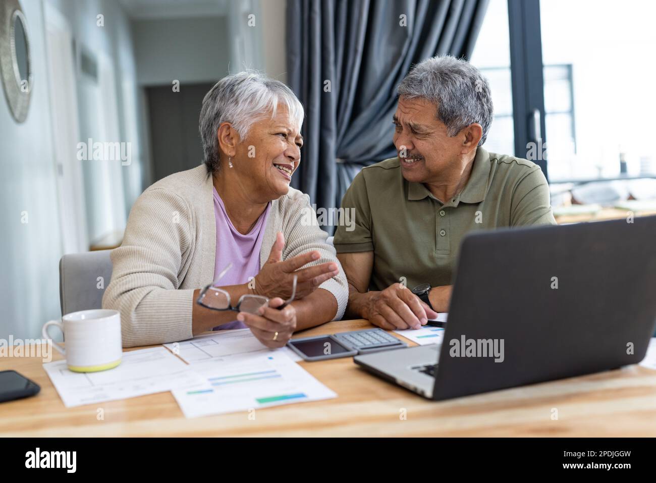 Biracial smiling senior couple with bills and laptop on table discussing finances at home Stock Photo