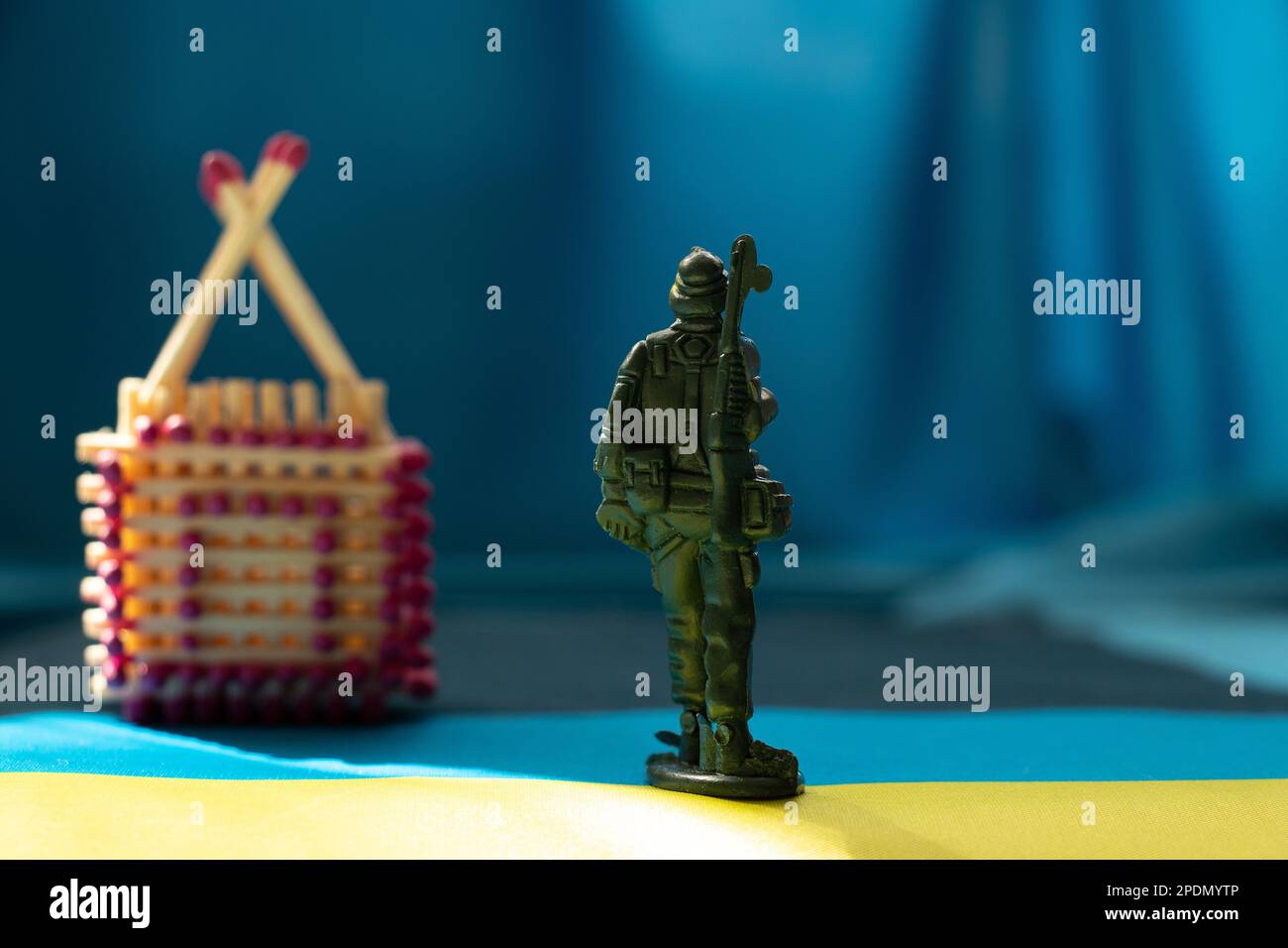 A small wooden house made of matches stands on the flags of Ukraine under the sun and a children's plastic soldier stands in front of the house, retur Stock Photo