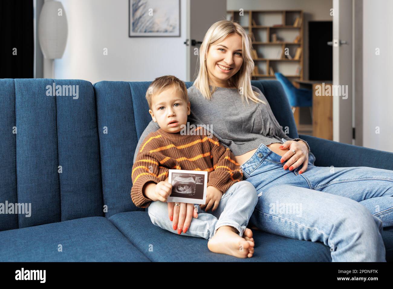 Young pregnant woman and her son sit on sofa at home and boy holds ultrasound scan in hand.They spend time together, enjoy communication feeling kicks Stock Photo