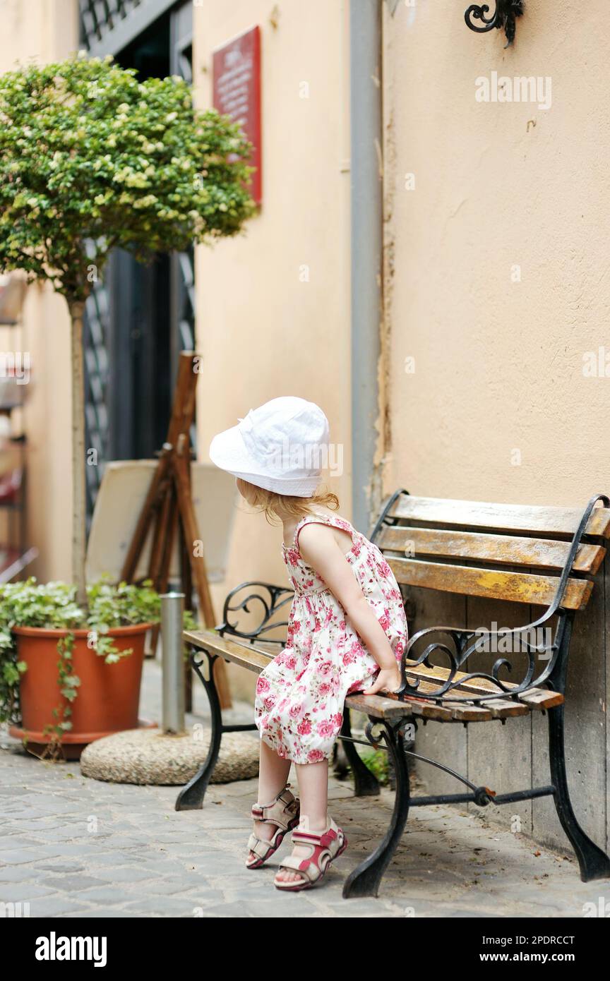 Cute little girl in floral dress sitting on a bench in beautiful town ...