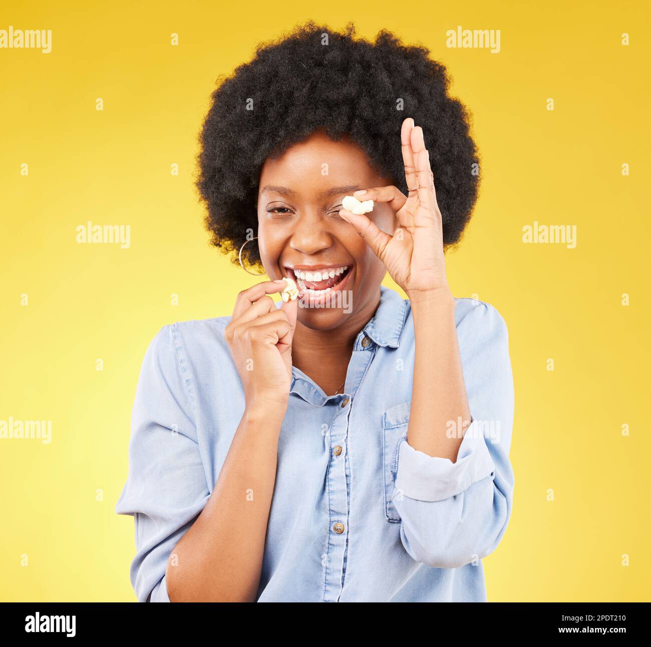 Black woman, funny face and eating popcorn in studio isolated on a yellow background. Comic smile, food and portrait of laughing, hungry and happy Stock Photo