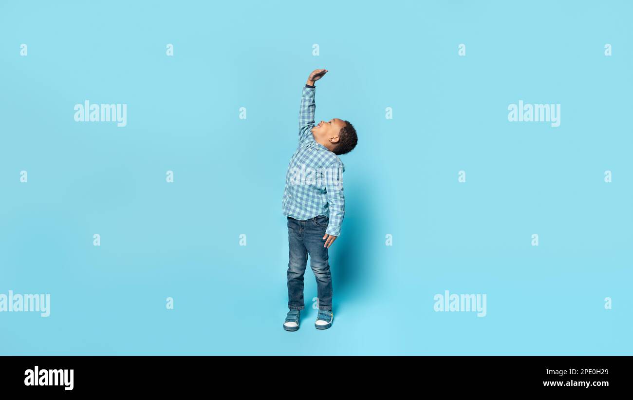 Little african american boy wishes he were taller, raising his hand to show how tall he wants to grow, blue background Stock Photo