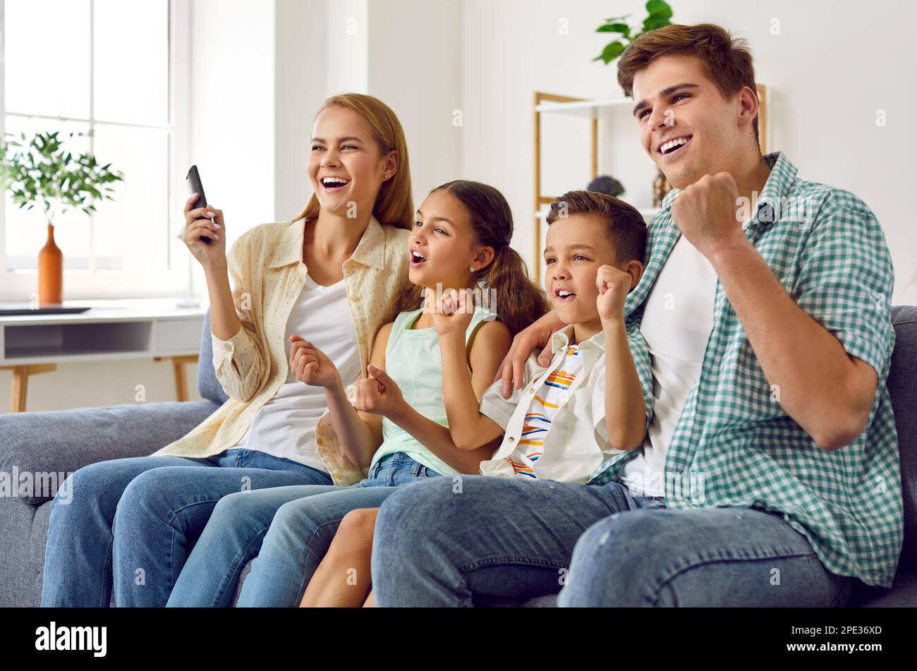 Cheerful family with children cheering together while watching TV at home in living room. Stock Photo