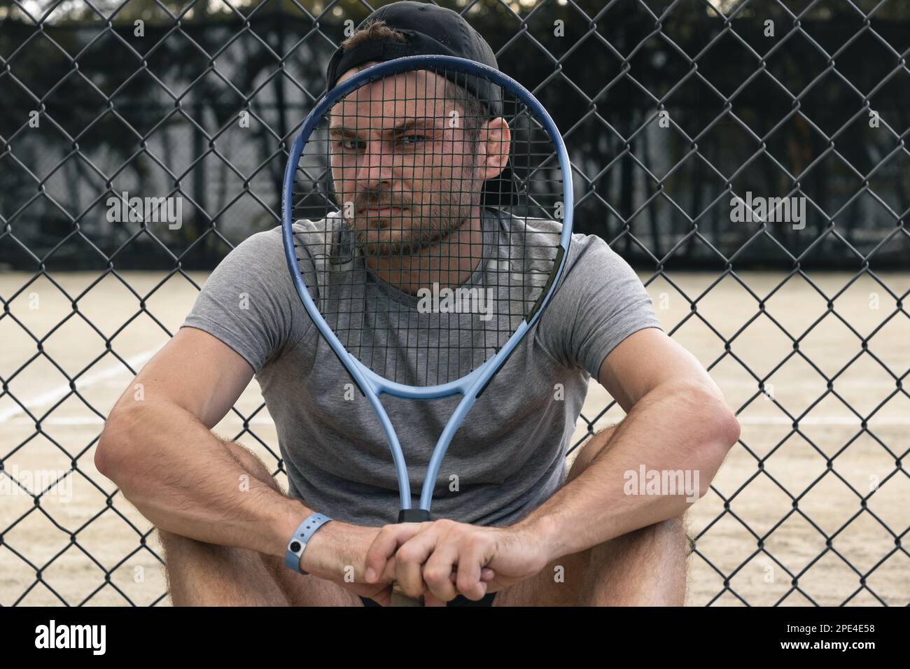 Tennis player sits on tennis court looking at camera through tennis racquet. Stock Photo