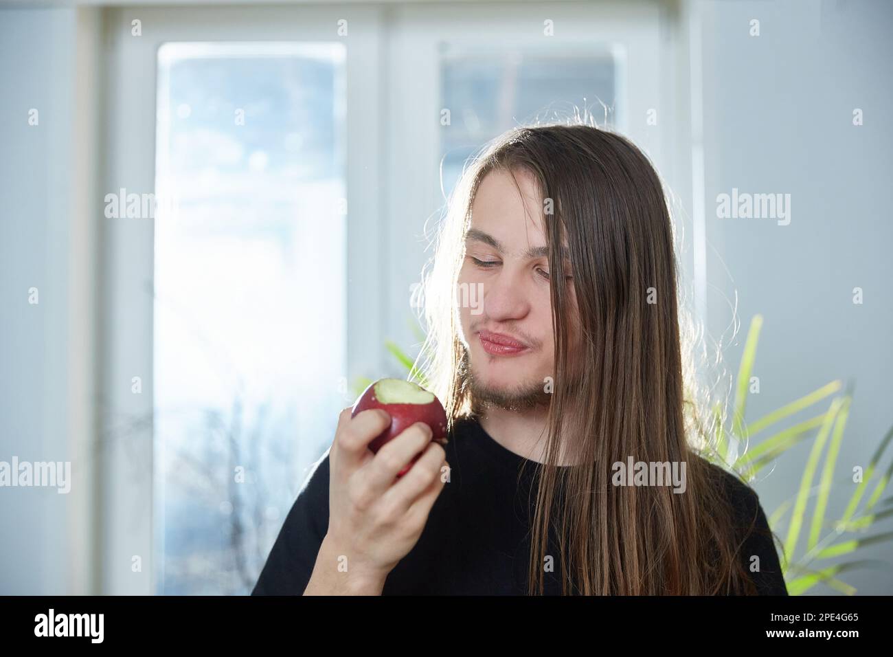 Hippie guy with long hair eats an apple at home. Stock Photo