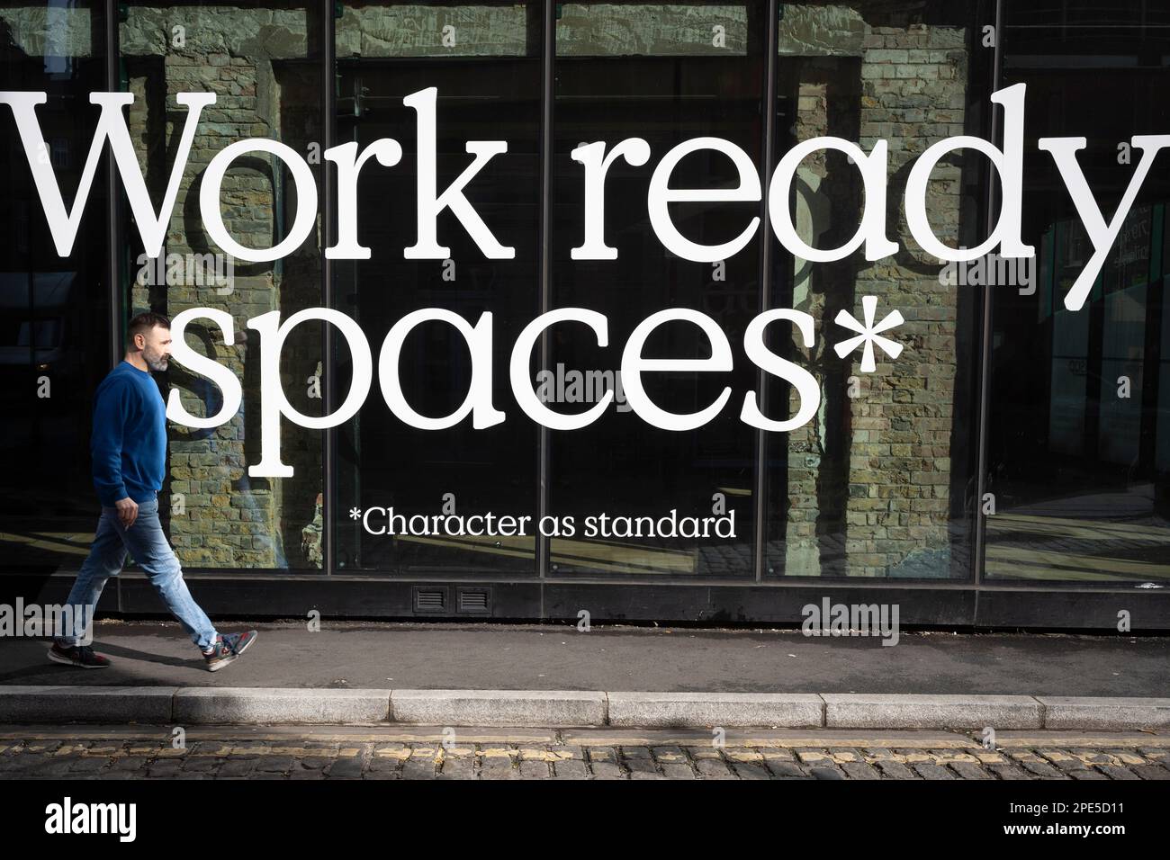 A bearded man strides past large lettering advertising work ready spaces in the window of a workplace property in Shoreditch, on 10th March 2023, in London, England. Stock Photo