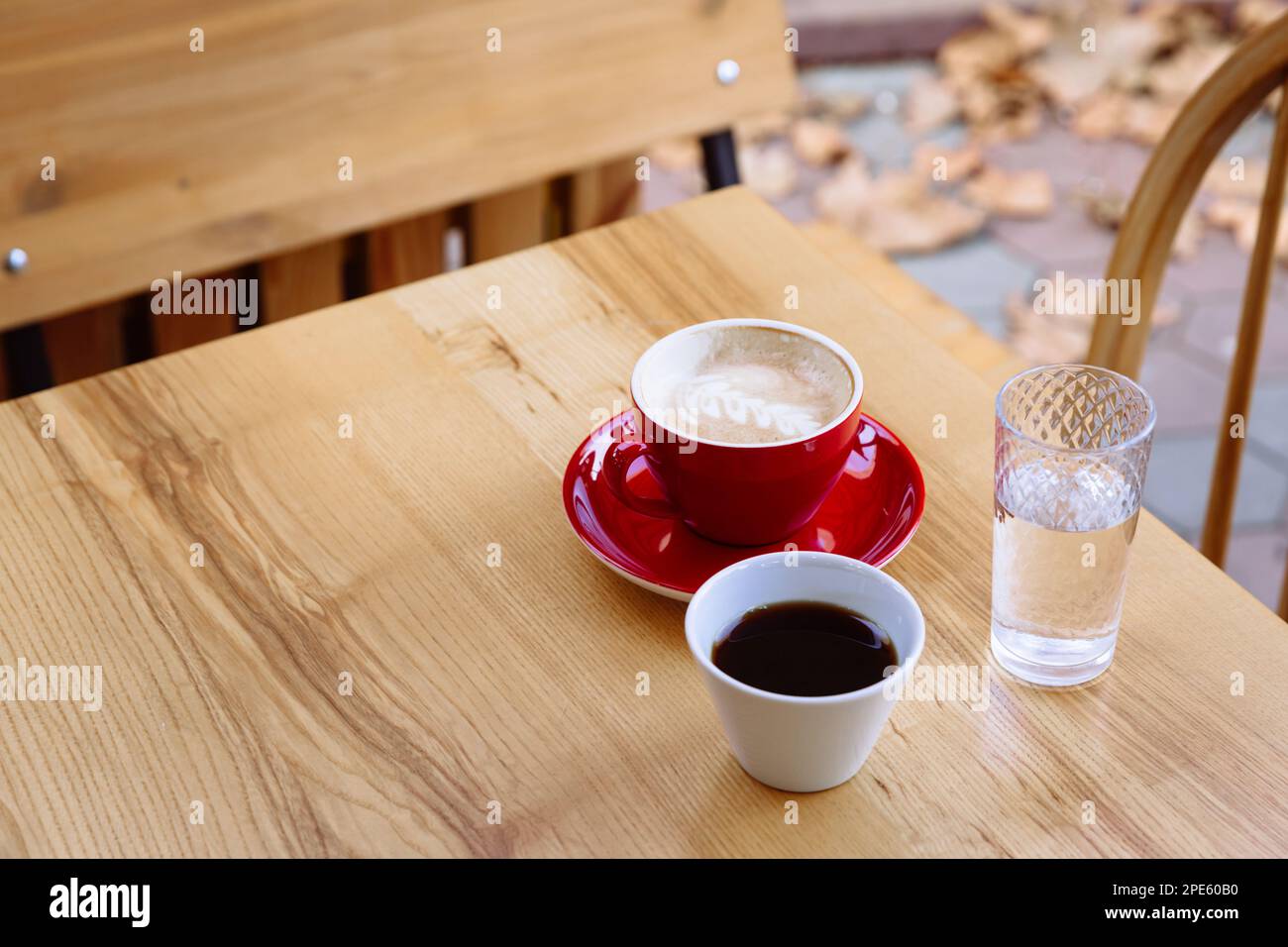 Aromatic coffee in a red cup with milk foam and latte art and freshly brewed coffee in a white cup. Nearby is a glass of water.Enjoyment of hot aroma beverage on fresh air. Stock Photo
