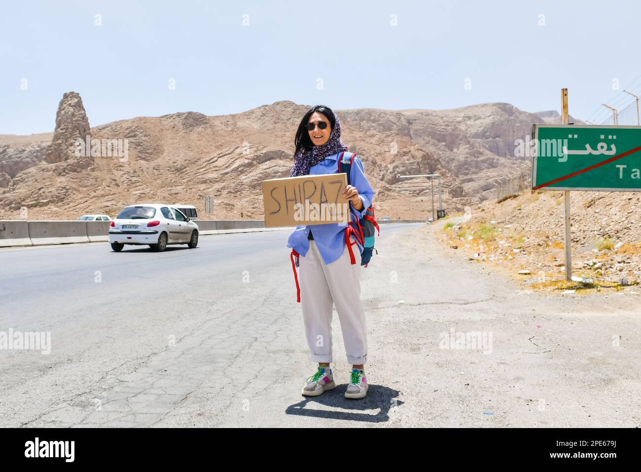 Young traveler woman stand on side of road hitchhike with sign to Shiraz and thumbs up with smile in Iran .Solo safe travel in Asia concept Stock Photo