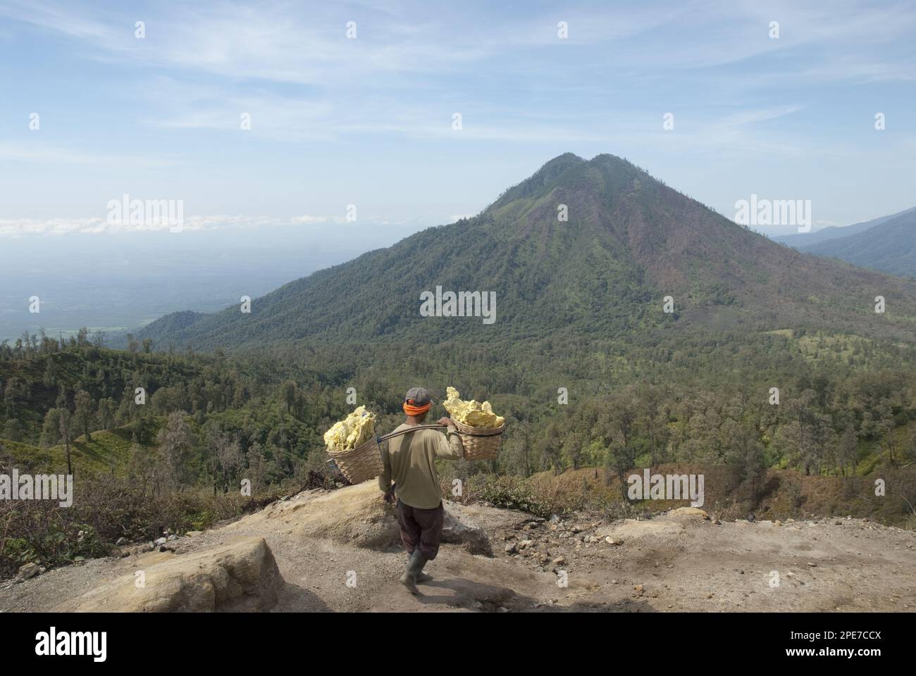 Local man carrying sulphur blocks in baskets down from the crater, with volcano in the background, Mount Ijen, East Java, Indonesia Stock Photo