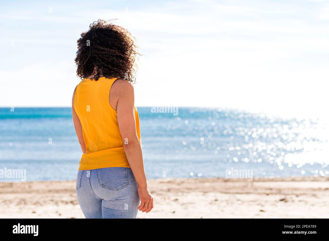 Anonymous curly haired female in yellow top and jeans standing on sandy beach while admiring waving sea during summer holidays Stock Photo