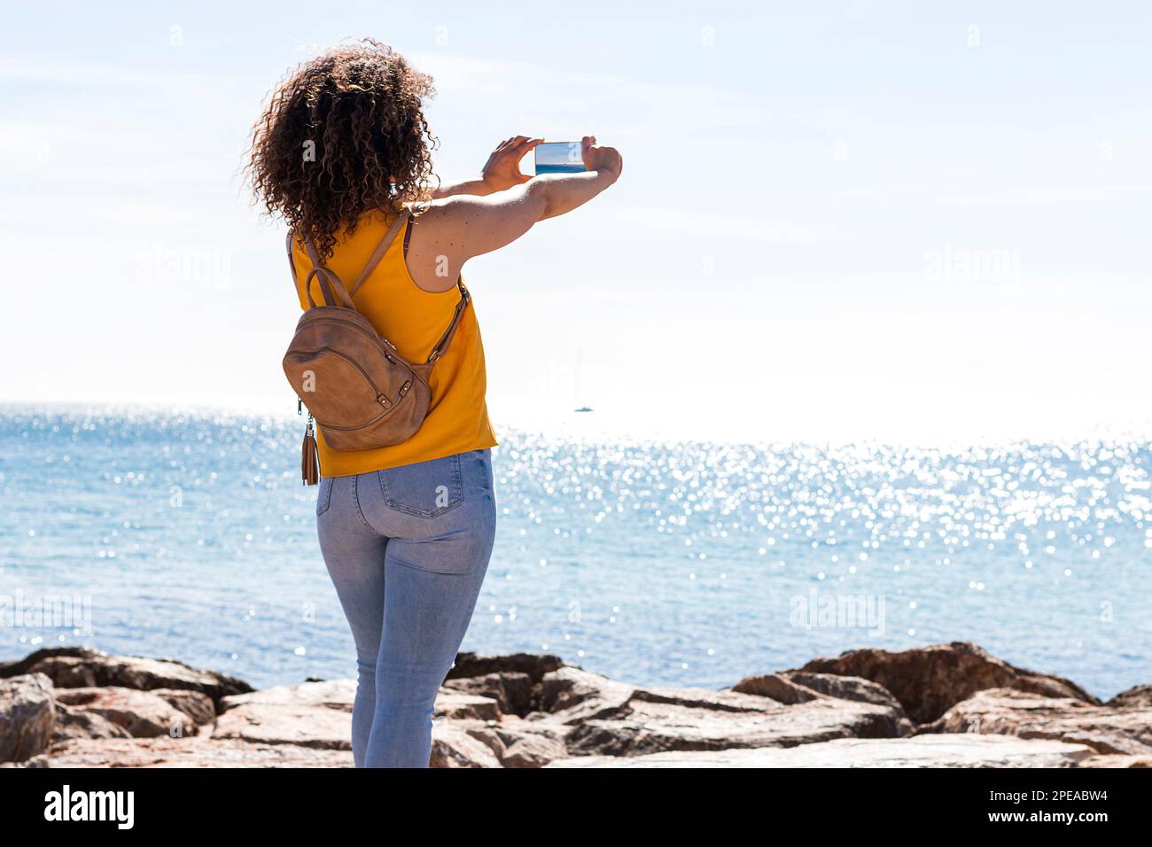 Unrecognizable curly haired female tourist in casual clothes with backpack standing on rocky coast and taking photo of waving sea on smartphone during Stock Photo