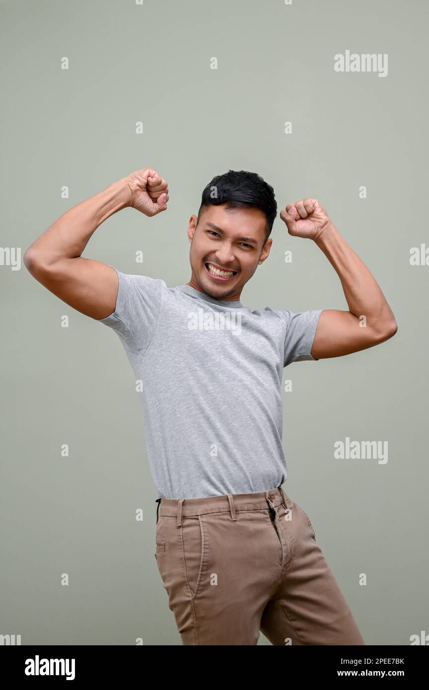 Happy and smiling millennial Asian man in casual clothes raising hand up, rejoicing, dancing with joy, yes gesture, winning, victory, celebrating, ach Stock Photo