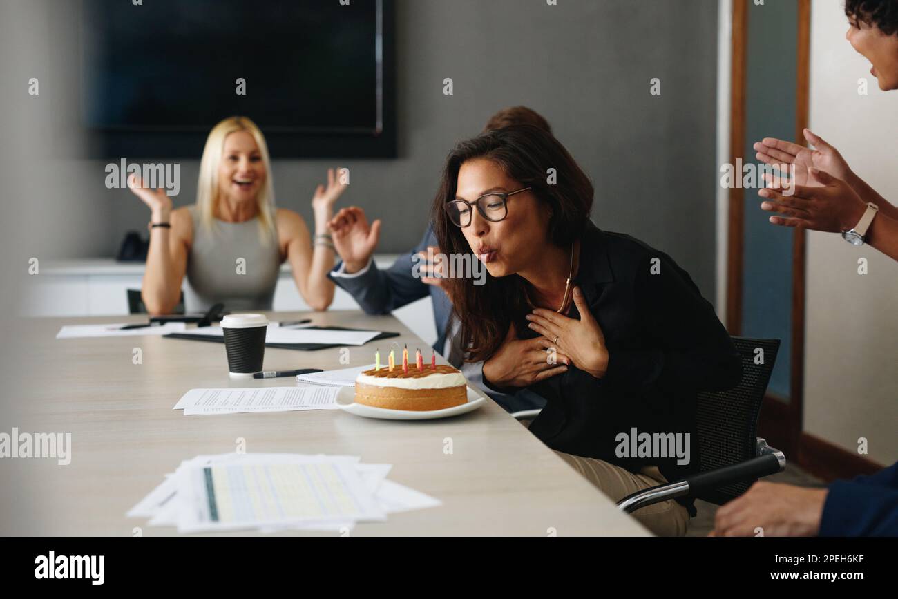 Young business woman celebrating her birthday at work with her colleagues. Woman blowing out candles on a cake at the office meeting table, with her c Stock Photo