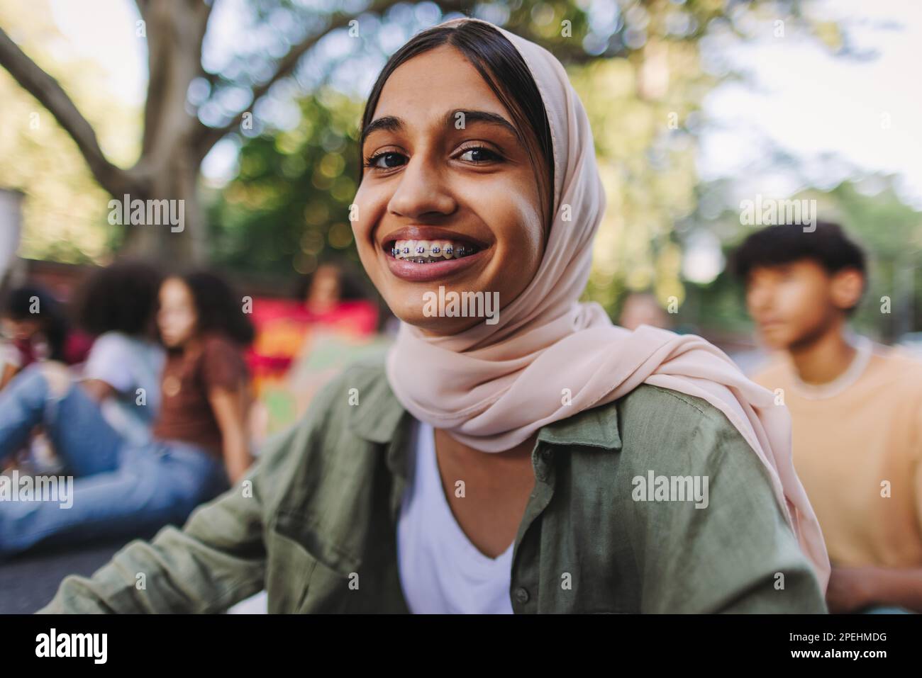 Cheerful Muslim girl smiling at the camera while sitting with a group of demonstrators at a climate protest. Multicultural youth activists joining the Stock Photo