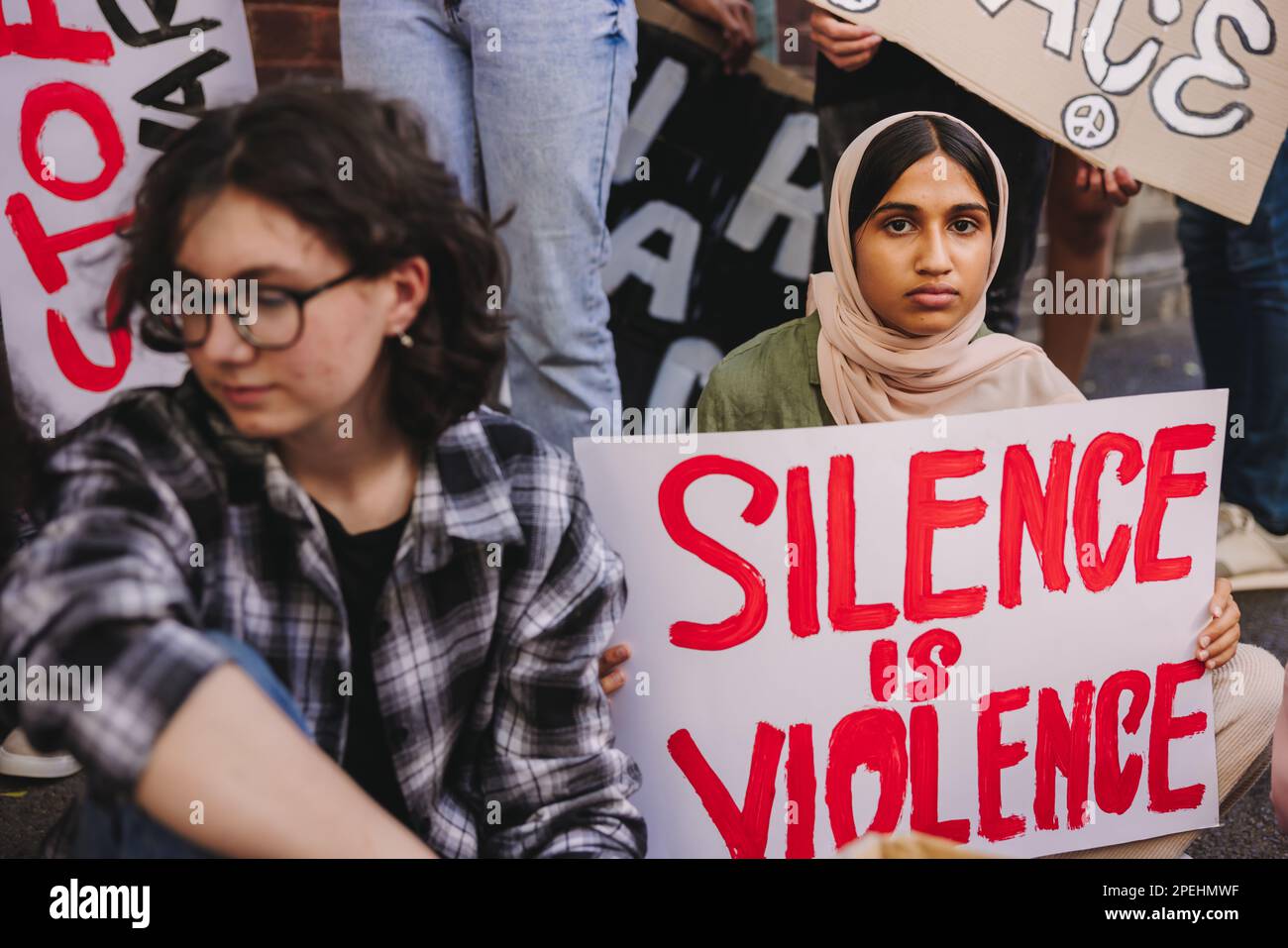 Young Muslim girl looking at the camera while holding an anti-violence poster. Diverse teenagers protesting against war and ethnic violence. Youth act Stock Photo