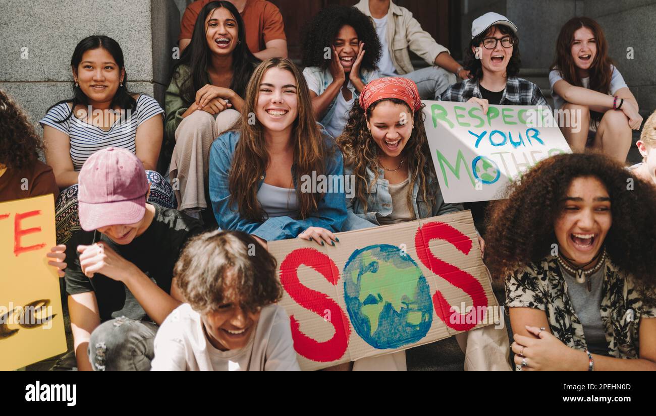 Youth environmental campaign. Group of multicultural climate activists smiling happily while sitting with posters and placards outside a building. You Stock Photo