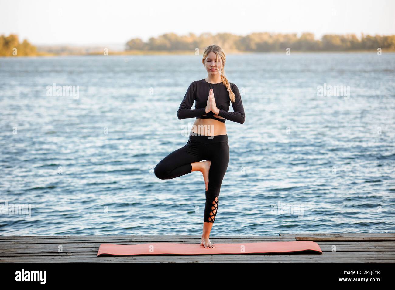 A young beautiful girl is doing yoga in a calm warm evening on the banks of the river. Stock Photo
