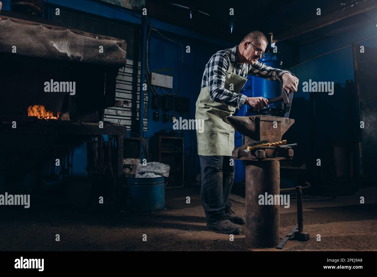 Senior an artisan blacksmith knocks with a hammer on iron to shape against the background of a burning forge. Stock Photo