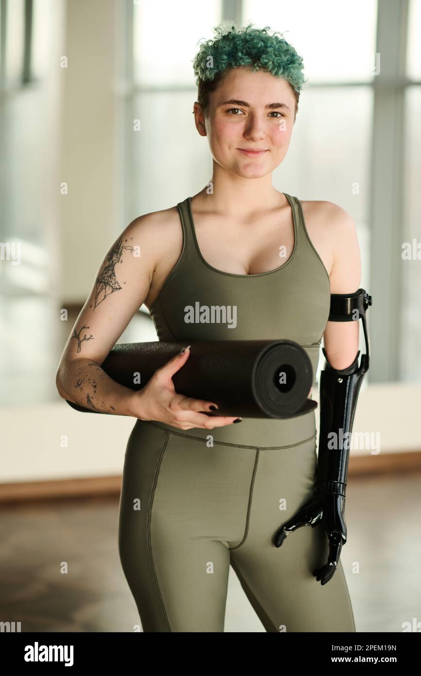 Portrait of young woman with prosthetic arm holding exercise mat and looking at camera while training in gym Stock Photo