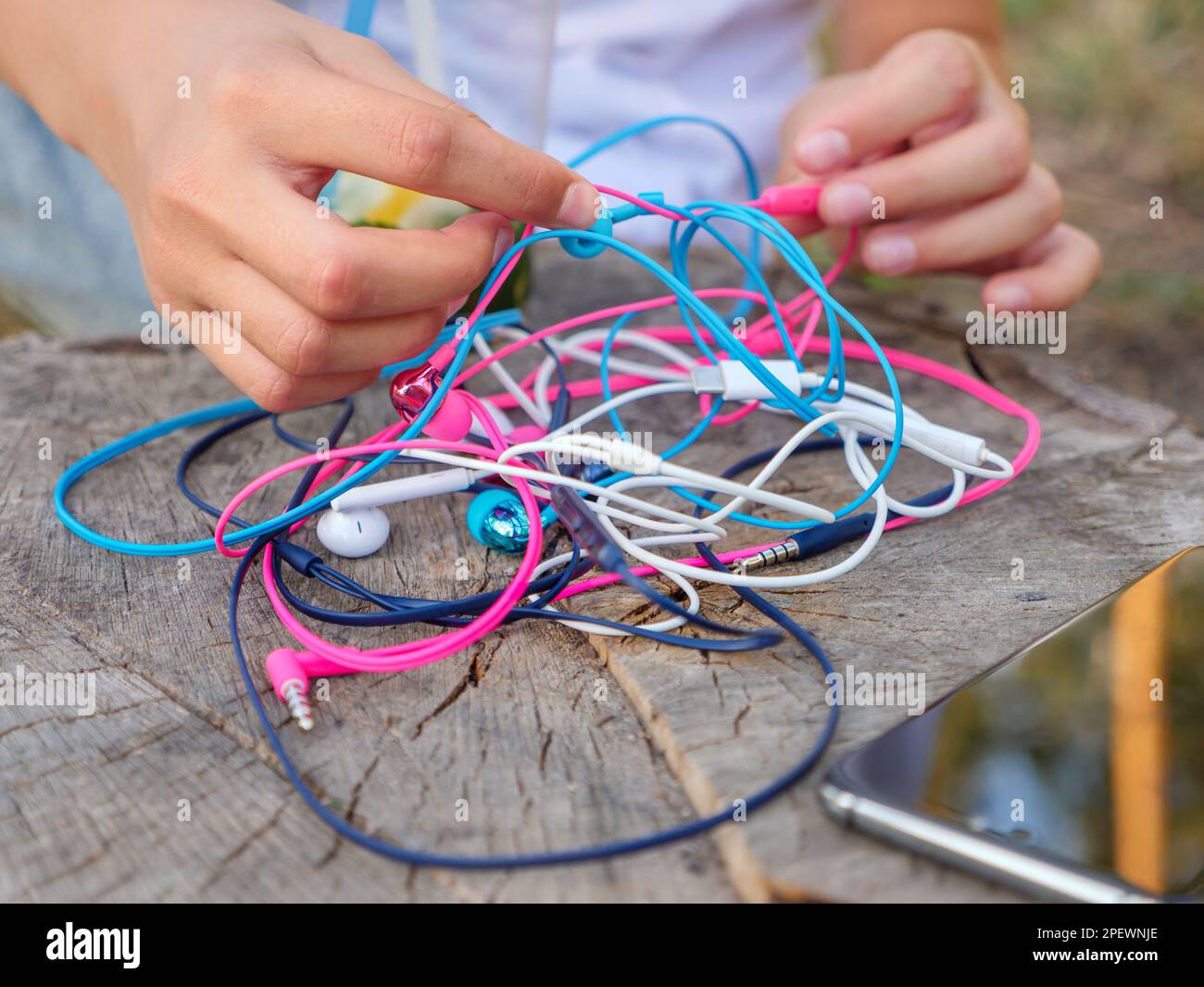 A phone and a hand holding a knot of four pairs of in-ear wired colorful pink, blue, white, and black earbuds tangled in a messy chaotic problem. Stock Photo