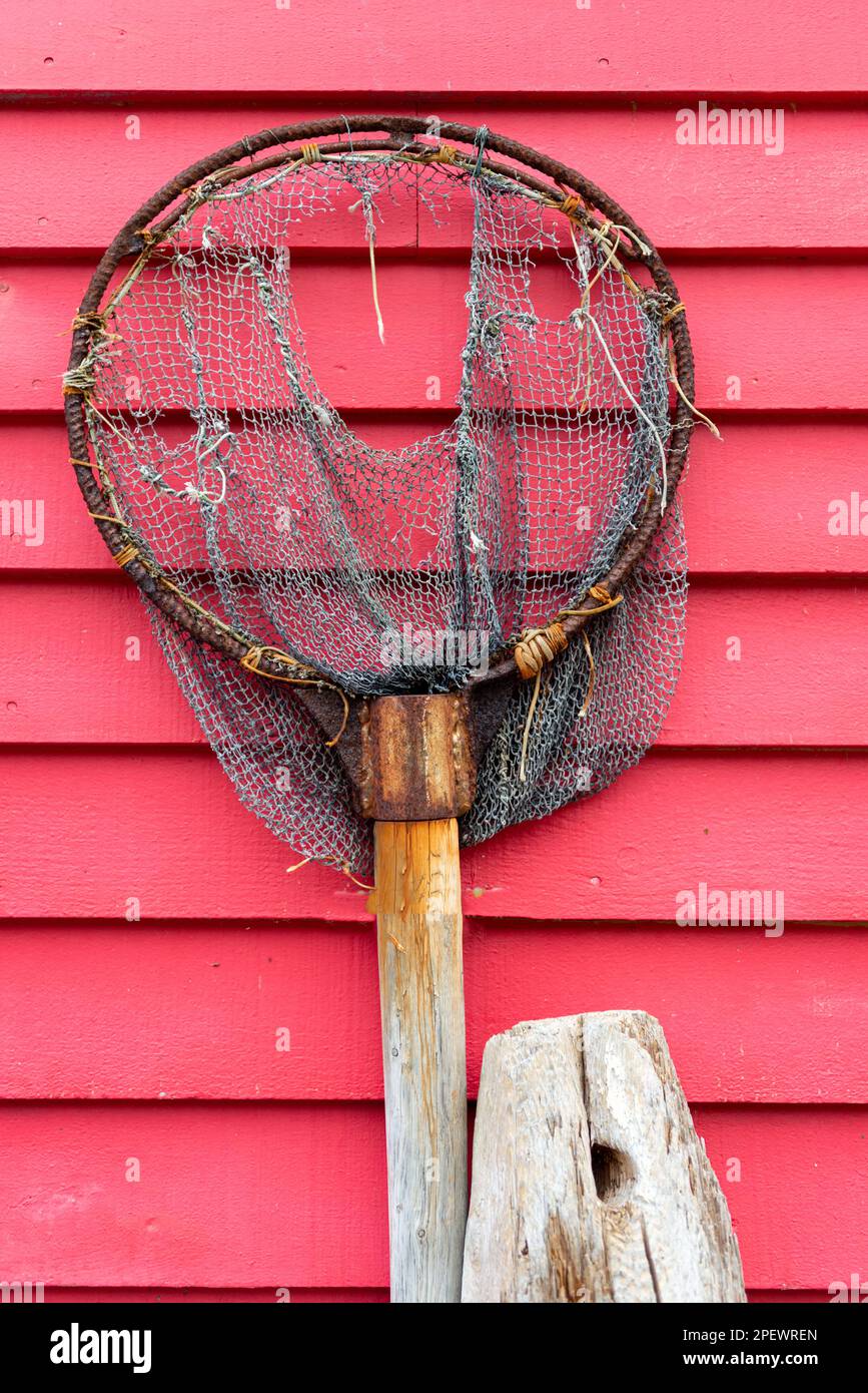 A vintage fishing dip net hanging on a vibrant red wooden horizontal clapboard wall. The scoop is rusty and the cloth mesh rope net is torn and worn. Stock Photo