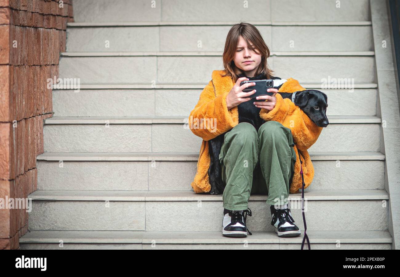 Young pretty woman in a yellow fur coat, social media influencer, sitting on the steps of the building city with her black dog, using smartphone. Stock Photo
