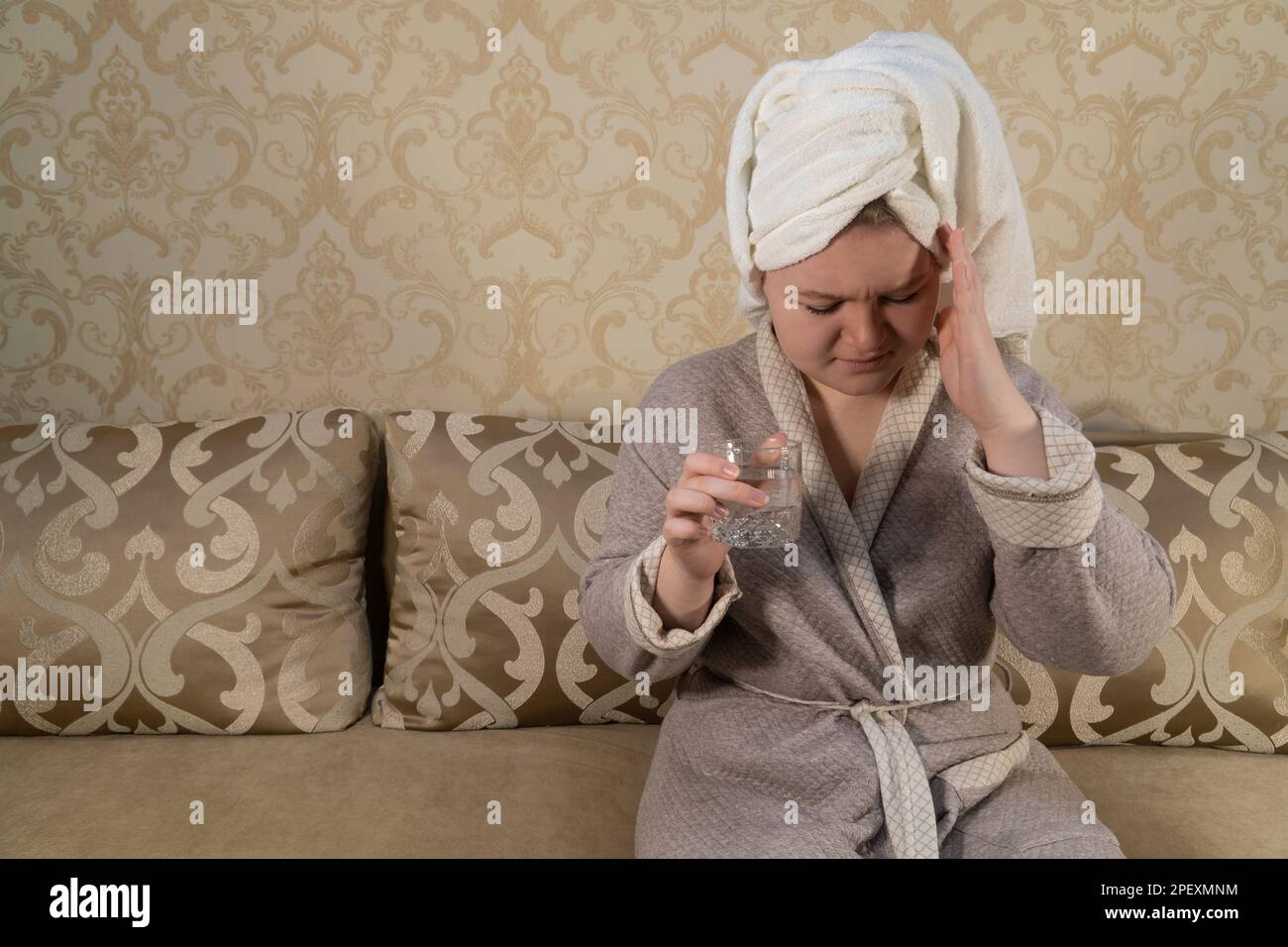 Girl in the room sits on the sofa and holds a glass of water in her hand. Headache. The concept of medicine, pharmacy, healthcare and home treatment Stock Photo