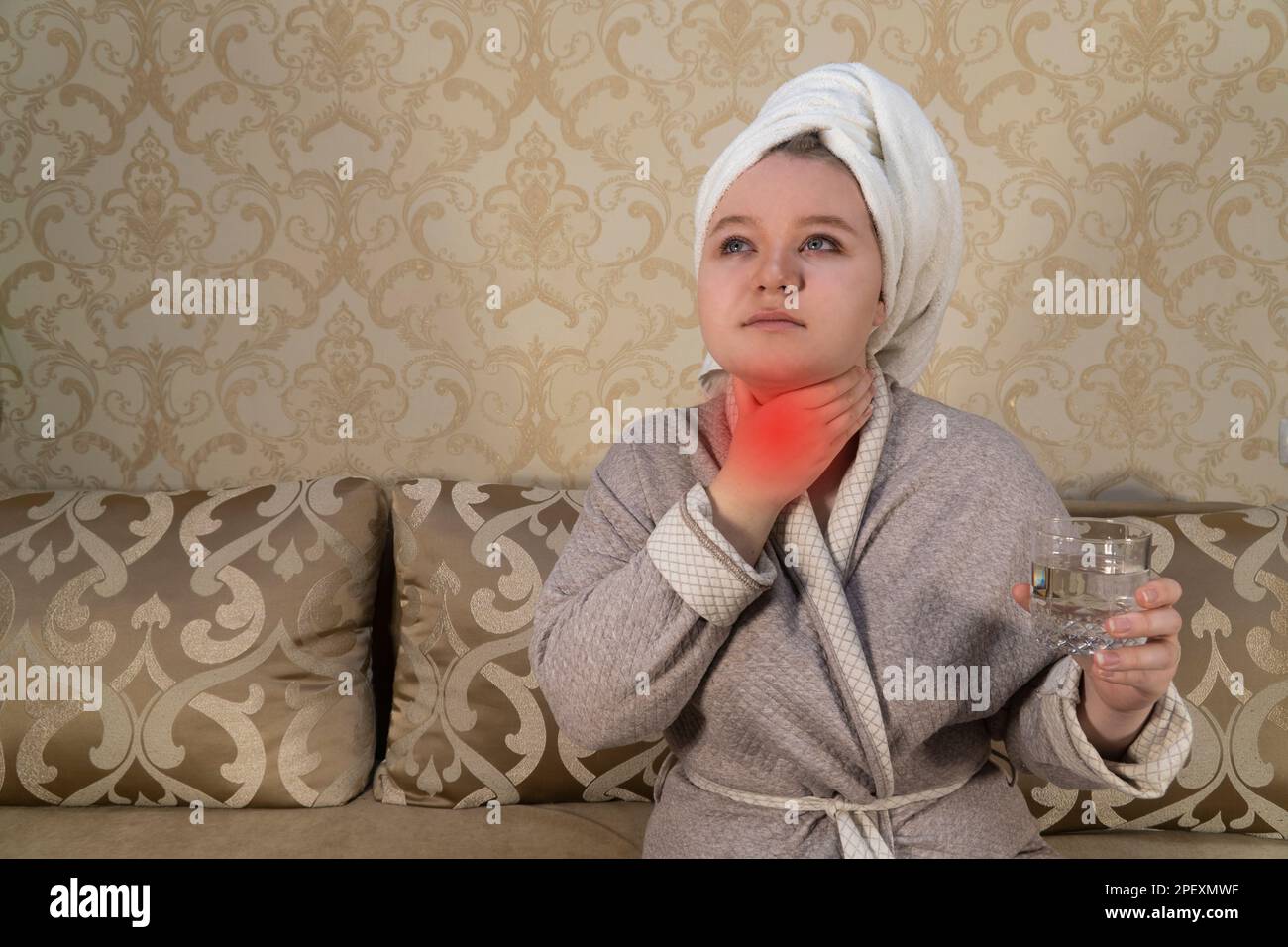Girl in the room sits on the couch and holds her throat in her hand. A sore throat. The concept of medicine, pharmacy, healthcare and home treatment Stock Photo