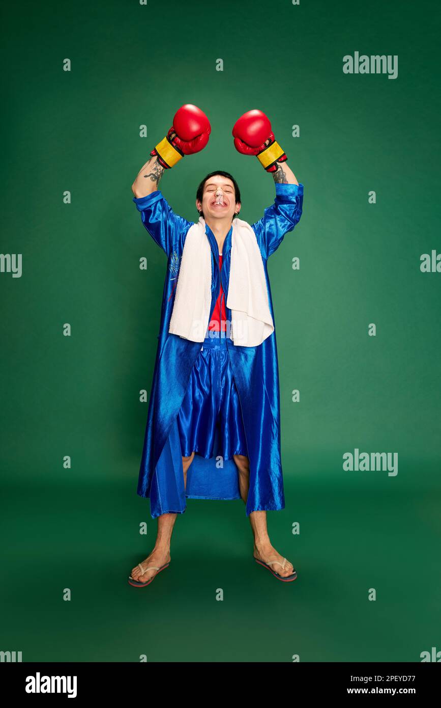 Portrait of boxer in uniform posing after win and keeping hands up above head isolated over green background. Concept of victory, sport, competition Stock Photo