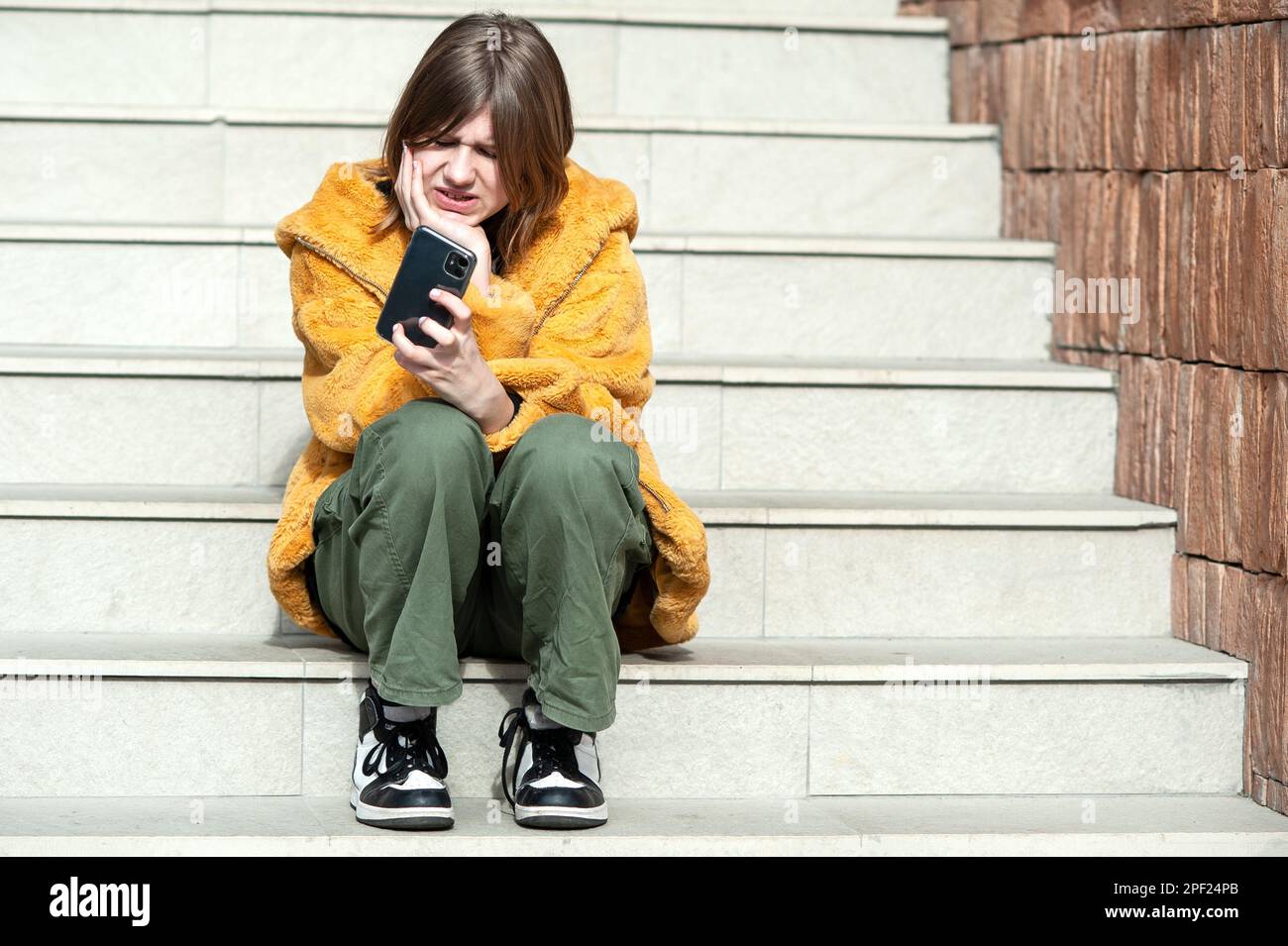 Young pretty woman in a yellow fur coat, social media influencer, sitting on stairs of the building city using smartphone. Stock Photo