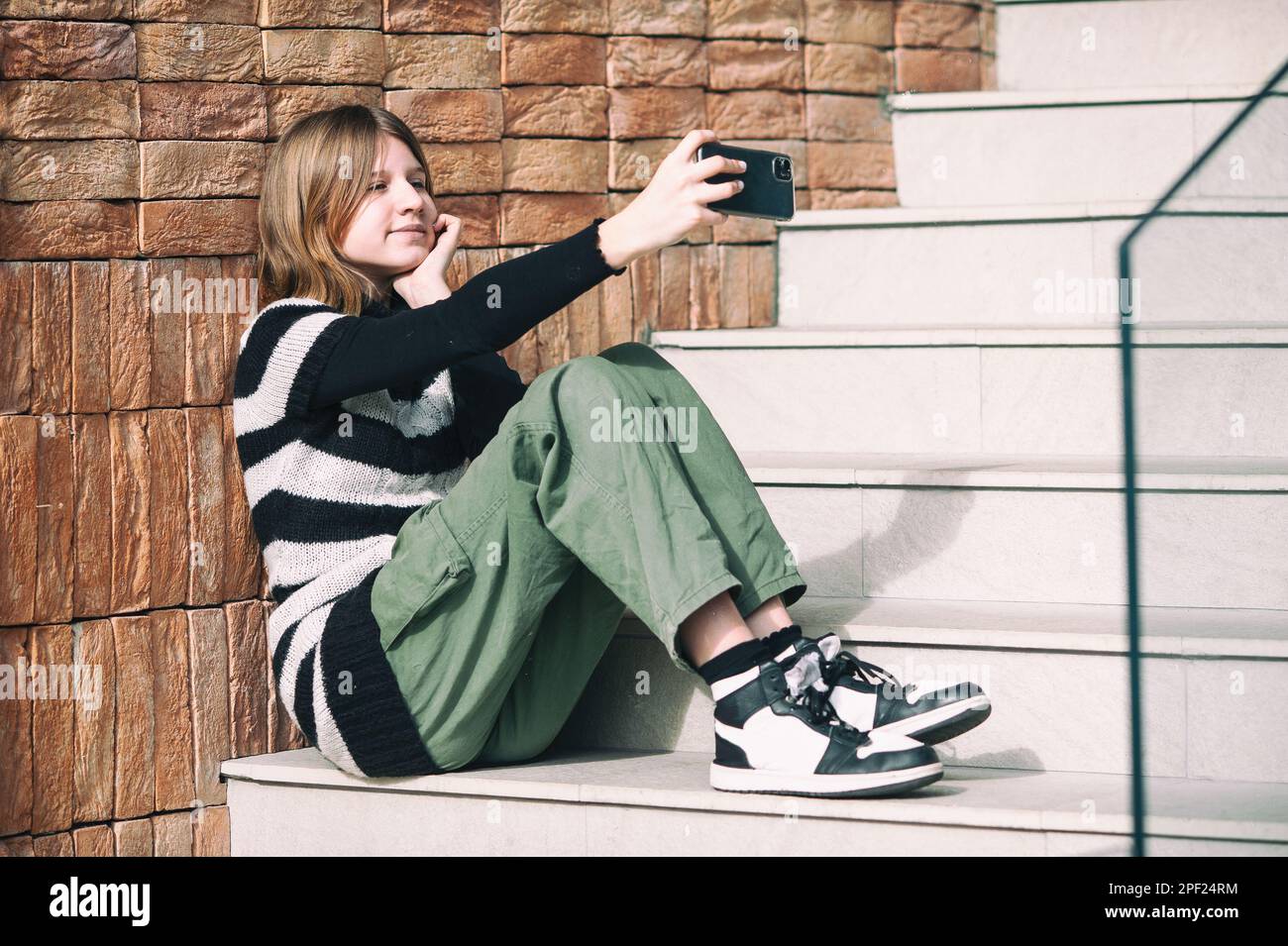 Young pretty woman in a striped black and white sweater, social media influencer, sitting on stairs of the building city using smartphone. Stock Photo
