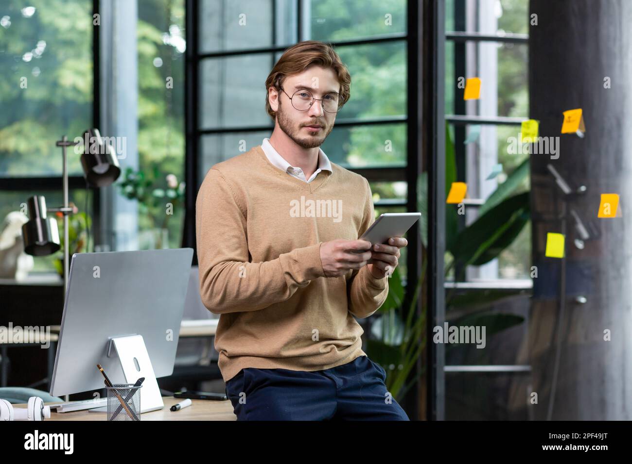 Portrait of a young male student, intern standing in the office, co-working at a table with a computer, using a tablet, studying online. Looks into the camera. Stock Photo