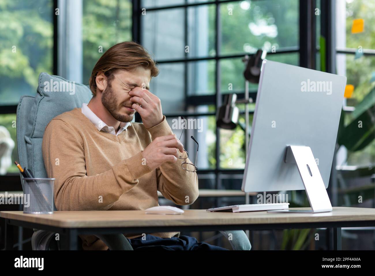 Tired male freelancer, programmer rubs his eyes, suffers from overfatigue, discomfort long sitting work in home office interior. Stock Photo