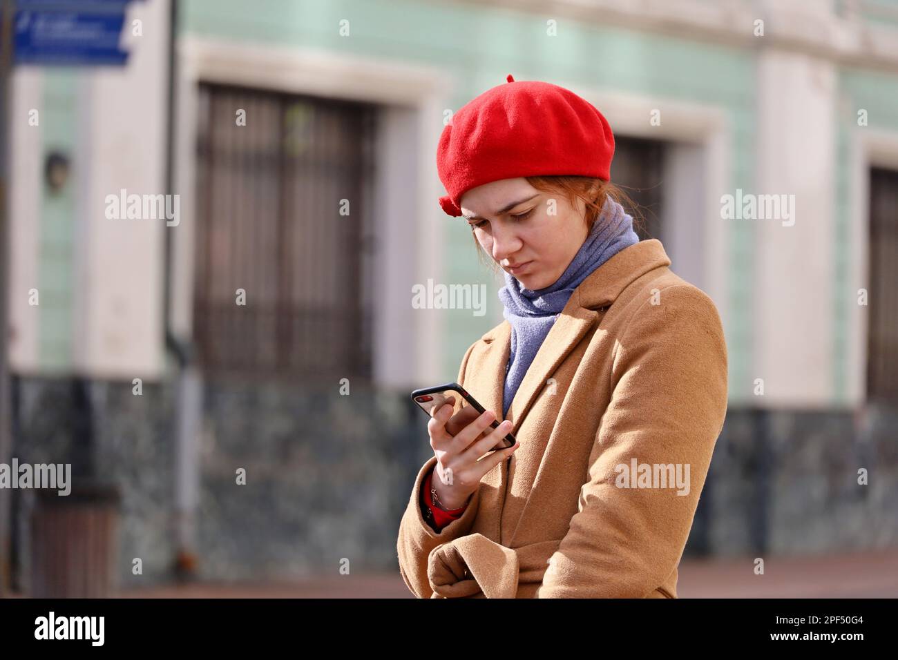 Pretty girl wearing coat and red cap standing with smartphone on a street. Using mobile phone in spring city Stock Photo