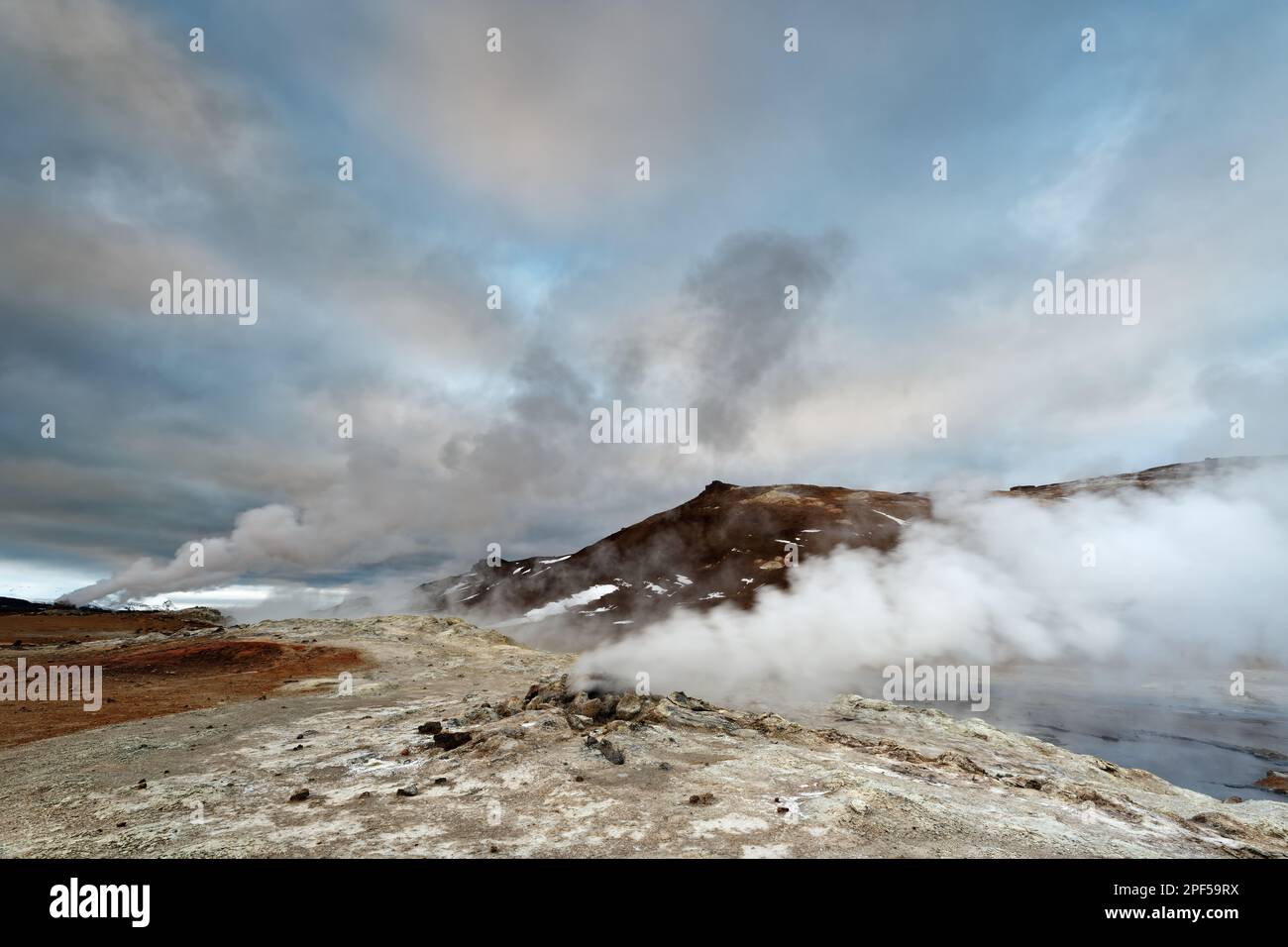 Wide volcanic landscape with steam escaping from fissures in the earth, blown sideways by the wind, above a sky with clouds coloured by the evening su Stock Photo