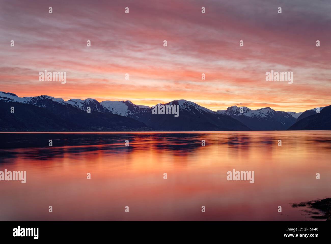 View over the water of a fjord in Norway in strong red evening light shortly after sunset, the mountains of the opposite shore appear in blue tones an Stock Photo