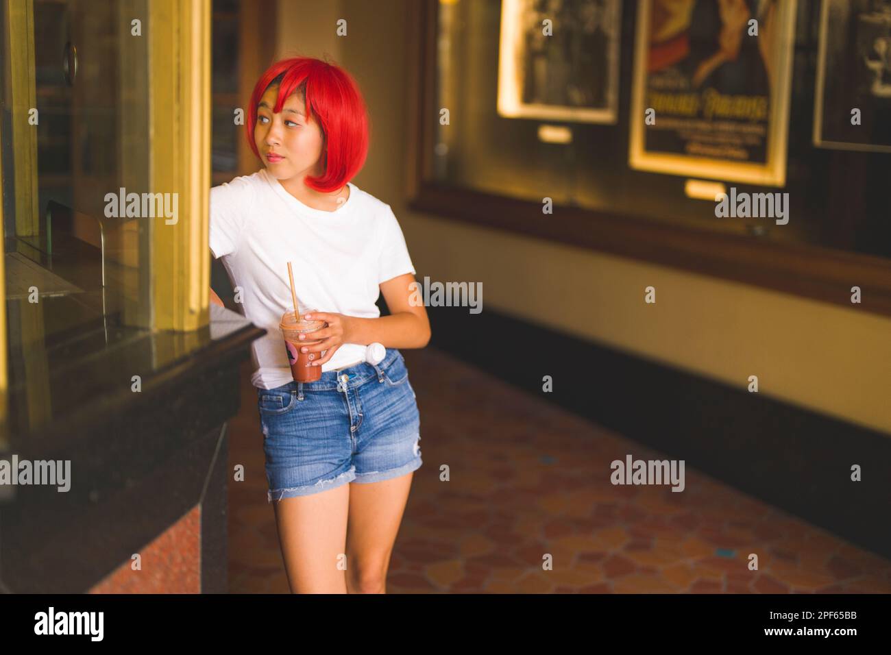 Modern Looking Teenage Girl in Bright Red Wig Leaning on Old Fashioned Movie Ticket Booth in Old Theater Stock Photo